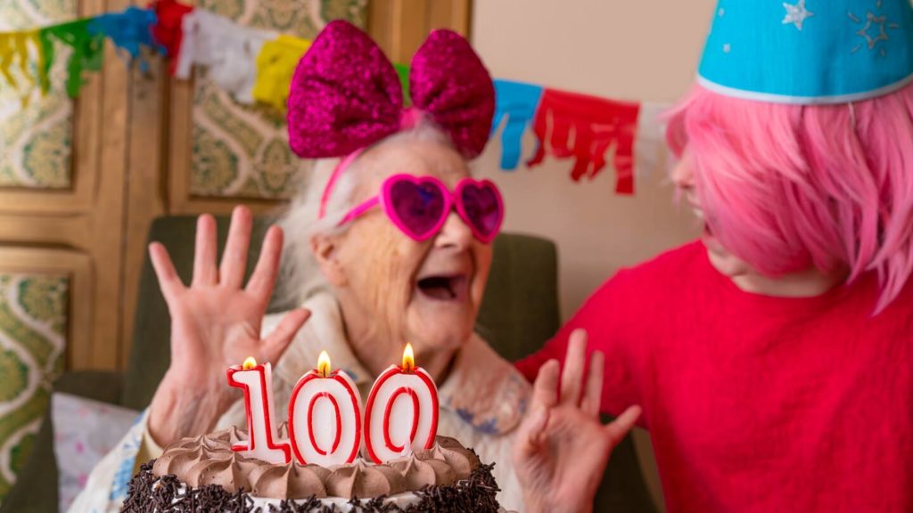 A centenarian celebrating her 100th birthday, wearing pink heart sunglasses and a giant purple bow on her head 